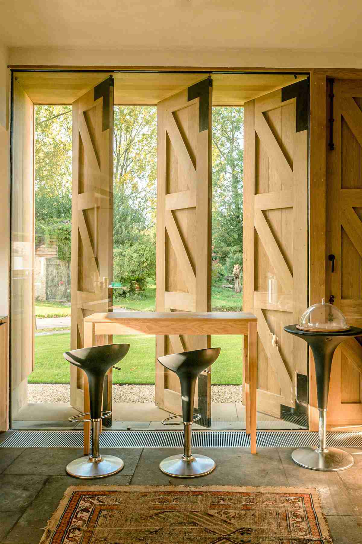 Custom timber bifold doors with vertical slats, paired with a wooden bar table and modern stools opening to a garden view
